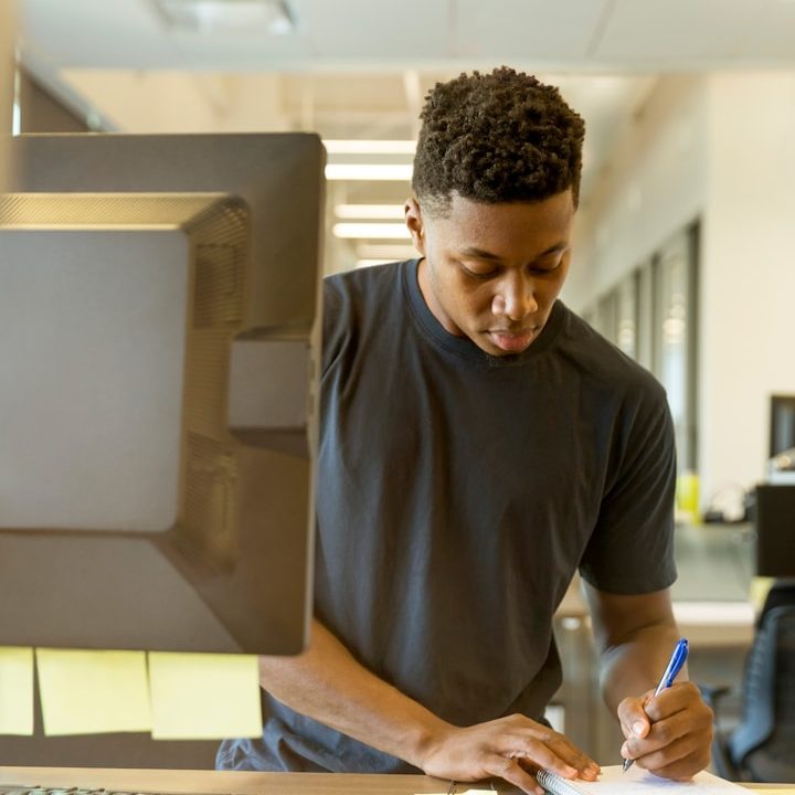 Small and Medium Size Business Man A man in a black t-shirt is writing notes at a desk with a computer.