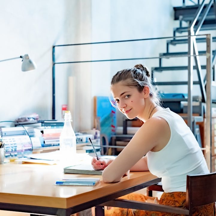 A freelancer and soletrader Young woman seated at a desk, writing in a notebook, with a bright light and staircase in the background.