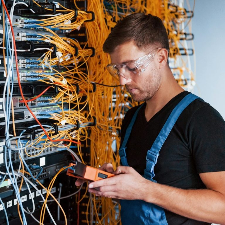 IT Solutions Technician in blue overalls checks equipment among tangled network cables.