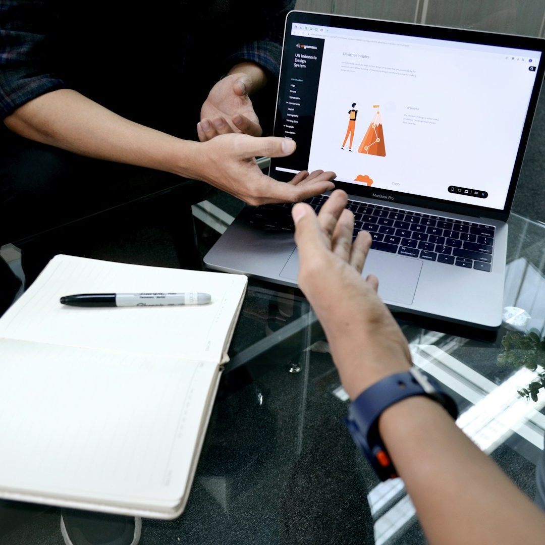 Picture for collaborative efforts Hands reaching towards a laptop displaying a design interface, with a notebook and pen nearby.